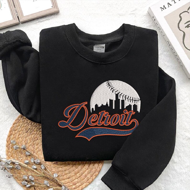 May include: Black sweatshirt with the word "Detroit" in orange script and a white baseball design. The sweatshirt has a crew neck and long sleeves. The design features a baseball with a skyline.