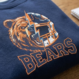 Puede incluir: Una sudadera azul marino con un bordado que muestra una cabeza de oso grizzly fusionada con un casco de f&uacute;tbol americano, dividida por un rayo. La palabra BEARS est&aacute; bordada debajo en letras de bloque naranjas con un contorno azul marino.