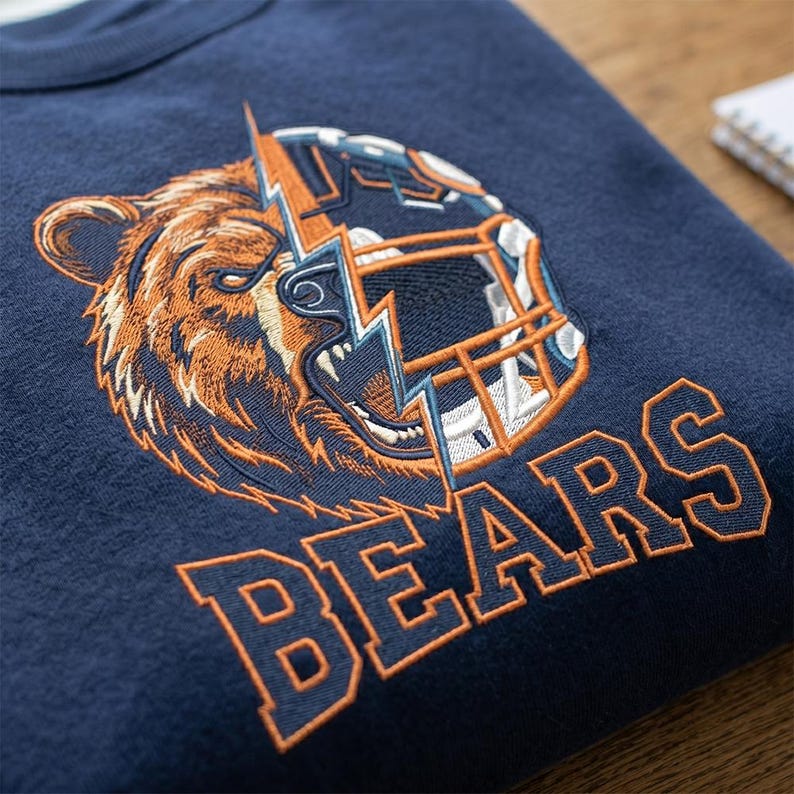 Puede incluir: Una sudadera azul marino con un bordado que muestra una cabeza de oso grizzly fusionada con un casco de f&uacute;tbol americano, dividida por un rayo. La palabra BEARS est&aacute; bordada debajo en letras de bloque naranjas con un contorno azul marino.