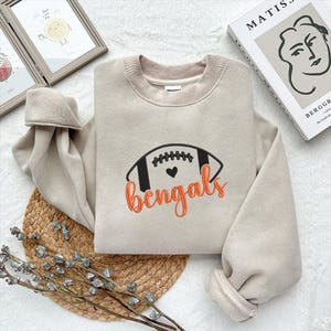 Puede incluir: Sudadera beige con un dise&ntilde;o de bal&oacute;n de f&uacute;tbol y la palabra "Bengals" en escritura naranja. El bal&oacute;n de f&uacute;tbol es negro con costuras blancas y un peque&ntilde;o coraz&oacute;n negro. La sudadera tiene cuello redondo y pu&ntilde;os acanalados.