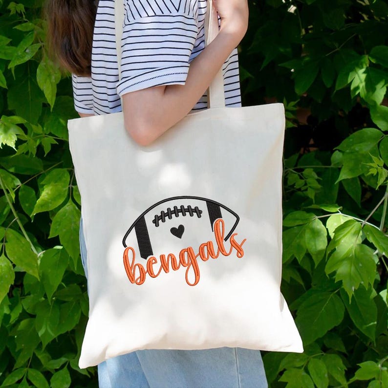 Puede incluir: Bolso tote color crema con un dise&ntilde;o de bal&oacute;n de f&uacute;tbol negro y naranja y la palabra "Bengals". El bolso tiene forma rectangular y correas largas. El dise&ntilde;o est&aacute; impreso en la parte delantera.