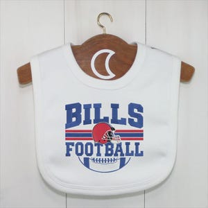 May include: White baby bib with the text "BILLS FOOTBALL" in blue, red, and white. The design includes a red football helmet and a football graphic. The bib is hanging on a wooden hanger.