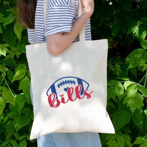May include: Cream-colored tote bag featuring a blue and red football design with the word "Bills" in red script. The bag has long, cream-colored handles and is held by a person wearing a striped shirt.