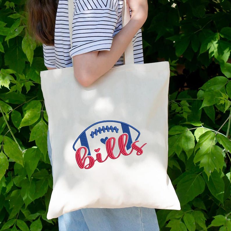 May include: Cream-colored tote bag featuring a blue and red football design with the word "Bills" in red script. The bag has long, cream-colored handles and is held by a person wearing a striped shirt.