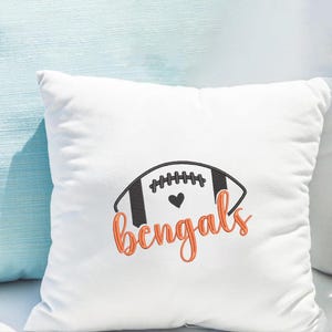 Puede incluir: Coj&iacute;n cuadrado blanco con un dise&ntilde;o de f&uacute;tbol americano bordado. El bal&oacute;n es negro y blanco con un coraz&oacute;n y la palabra "Bengals" en naranja. Art&iacute;culo decorativo.