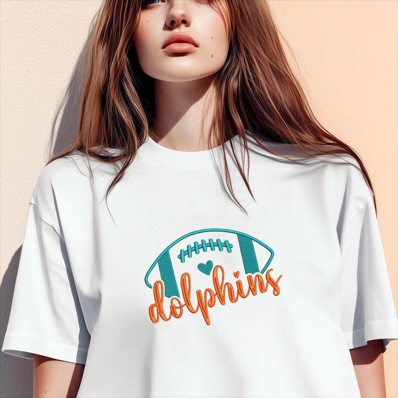 Puede incluir: Camiseta blanca con un dise&ntilde;o de f&uacute;tbol americano turquesa y naranja y la palabra "dolphins" en escritura naranja. El f&uacute;tbol americano tiene rayas turquesas y un coraz&oacute;n. Un look casual y deportivo.