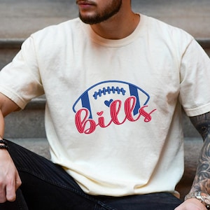 May include: Cream-colored t-shirt featuring a football design with the word "Bills" in red script. The football is blue with white stitching and a heart detail. A casual, sporty apparel item.