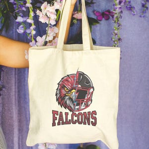 Puede incluir: Una bolsa de tela color crema con un dise&ntilde;o gr&aacute;fico dividido que muestra la cabeza de un halc&oacute;n a la izquierda y un casco de f&uacute;tbol americano a la derecha, separados por un rayo. La palabra FALCONS est&aacute; impresa en rojo oscuro bajo el gr&aacute;fico.