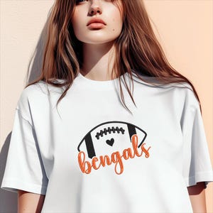 Puede incluir: Camiseta blanca con un dise&ntilde;o de f&uacute;tbol americano negro y naranja y la palabra "Bengals" en escritura naranja. El f&uacute;tbol americano tiene rayas negras y un coraz&oacute;n. Una prenda casual y deportiva.