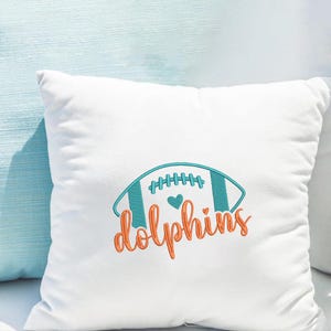Puede incluir: Coj&iacute;n cuadrado blanco con un dise&ntilde;o de f&uacute;tbol bordado en verde azulado y naranja. El dise&ntilde;o incluye la palabra "Dolphins" en una fuente cursiva y un peque&ntilde;o coraz&oacute;n.