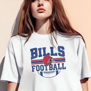 May include: White t-shirt featuring the text "BILLS FOOTBALL" in blue, with a red football helmet graphic. The design includes red and blue stripes and a football graphic. A casual, sporty apparel item.
