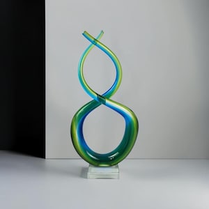 May include: A tall, abstract glass sculpture featuring a continuous, flowing form that twists into an infinity-like shape. The piece is crafted from translucent glass with vibrant gradients of emerald green and deep ocean blue, mounted on a clear base.