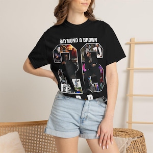 Pu&ograve; includere: T-shirt nero con la scritta "RAYMOND & BROWN" sopra il numero "26". Il numero &egrave; riempito con un collage di immagini. La modella indossa pantaloncini di jeans azzurri.