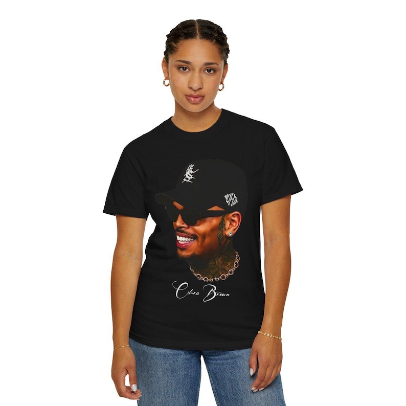Pu&ograve; includere: T-shirt nero con un ritratto di un uomo sorridente con berretto da baseball e occhiali da sole. L'immagine include il testo "Chris Brown" in corsivo. La maglietta &egrave; indossata da una persona.