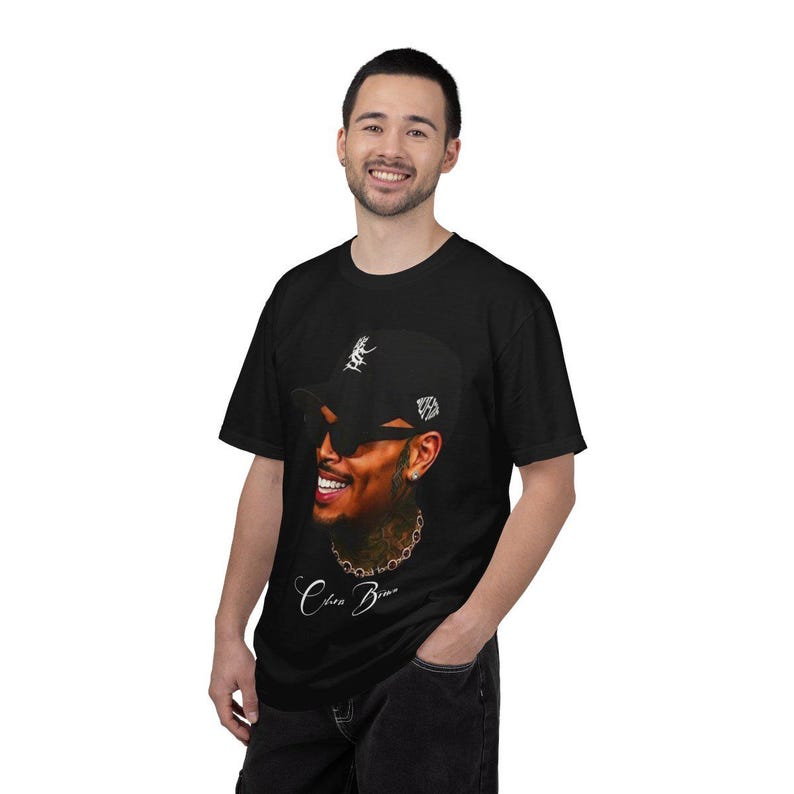 Pu&ograve; includere: T-shirt nero con un ritratto di una persona sorridente che indossa un berretto da baseball nero e occhiali da sole. Il berretto ha un logo bianco. La maglietta ha il nome "Chris Brown" in corsivo.