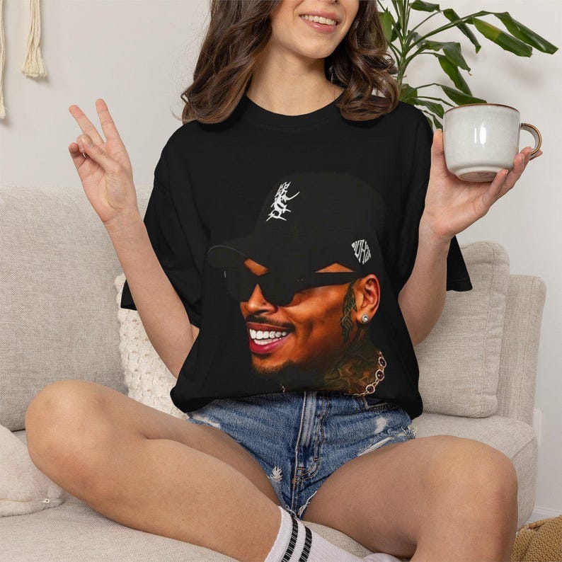 Pu&ograve; includere: T-shirt nero con un ritratto sorridente di un uomo con berretto nero e occhiali da sole. Il berretto ha disegni grafici bianchi. La persona nell'immagine indossa una catena d'oro. La persona indossa pantaloncini di jeans.