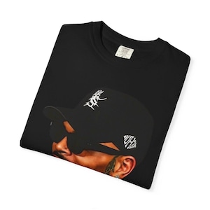 Pu&ograve; includere: T-shirt nero girocollo con grafica di una persona con berretto nero e occhiali da sole. Il berretto ha scritte bianche. La maglietta ha un'etichetta con la scritta "Comfort Colors".