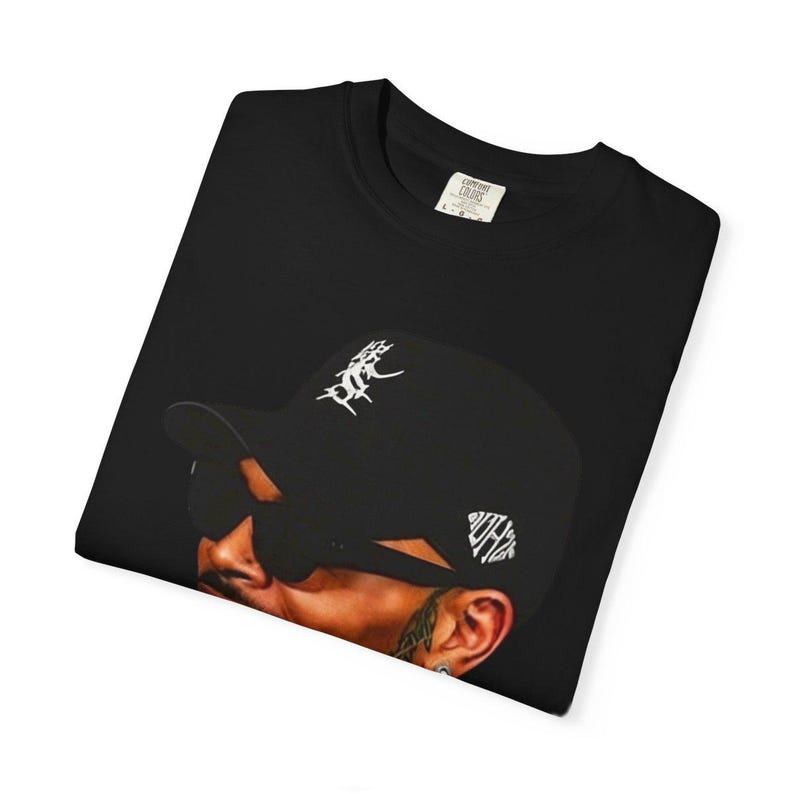 Pu&ograve; includere: T-shirt nero girocollo con grafica di una persona con berretto nero e occhiali da sole. Il berretto ha scritte bianche. La maglietta ha un'etichetta con la scritta "Comfort Colors".