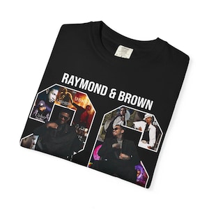 Pu&ograve; includere: T-shirt nero con la scritta bianca "RAYMOND & BROWN". La maglietta presenta un collage di immagini di musicisti. La maglietta &egrave; piegata su una superficie bianca.