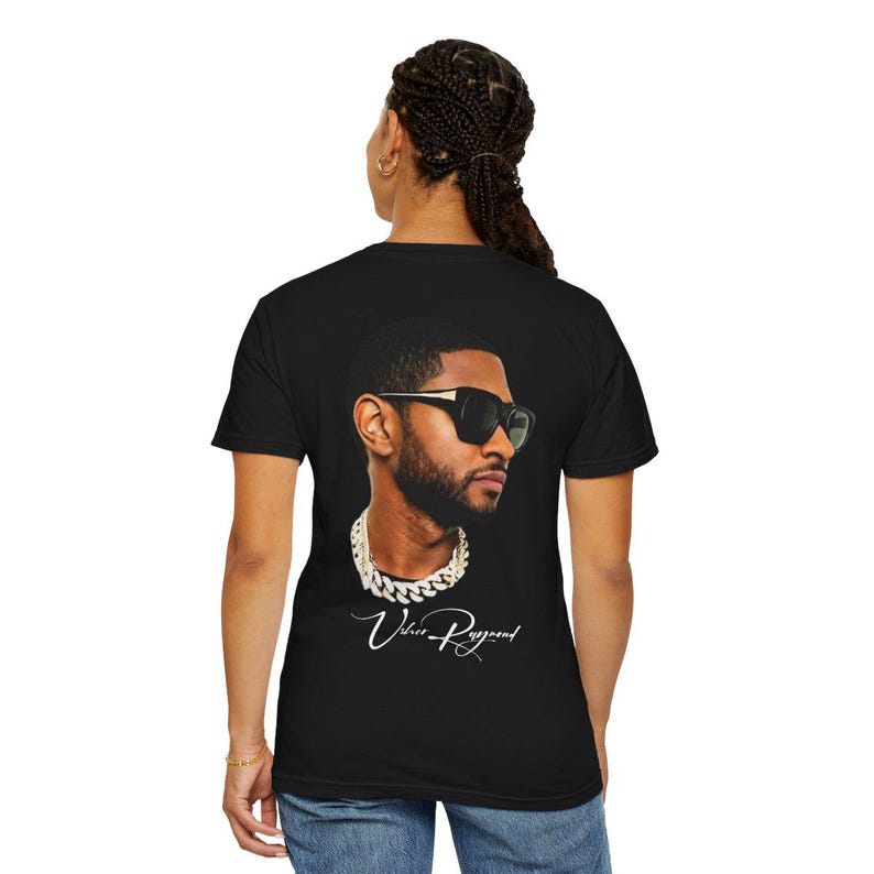 Pu&ograve; includere: T-shirt nero con un ritratto di Usher con occhiali da sole e collana a catena. L'immagine include il testo "Usher Raymond" in corsivo. La maglietta &egrave; un classico girocollo.