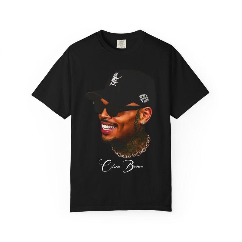Pu&ograve; includere: T-shirt nero con un ritratto sorridente di Chris Brown con occhiali da sole e berretto da baseball. Il testo "Chris Brown" &egrave; scritto in corsivo sotto. La maglietta &egrave; a girocollo.
