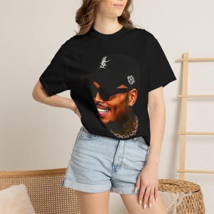 Pu&ograve; includere: T-shirt nero con un ritratto di una persona sorridente con un berretto nero e occhiali da sole. Il berretto ha un testo bianco e un disegno. La persona indossa una catena d'oro. La modella indossa pantaloncini di jeans blu.