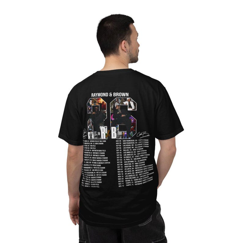Pu&ograve; includere: T-shirt nera con la scritta "RAYMOND & BROWN" sopra una grafica di due persone. La grafica include il numero "23" e un elenco di date e luoghi del tour. Una firma &egrave; visibile sul lato destro della grafica.
