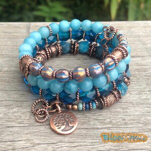 May include: Stackable beaded bracelets in shades of blue and copper. The bracelets feature various bead sizes and shapes, including round and faceted beads. A copper charm with a tree design hangs from one bracelet. The bracelets are displayed on a wooden surface.