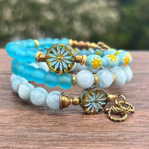 Könnte beinhalten: Drei Armbänder mit blauen und goldenen Perlen. Das obere Armband hat durchscheinende blaue Perlen, das mittlere hat mattierte blaue Perlen und das untere hellblaue Perlen. Jedes Armband hat dekorative goldene Akzente und Perlen mit Blumenmuster.