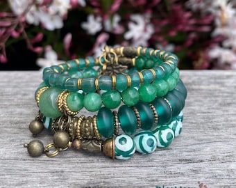 May include: A collection of green beaded bracelets with gold-tone details. The bracelets showcase a variety of bead shapes and textures, including round, cylindrical, and patterned beads. The bracelets are presented on a wooden surface.