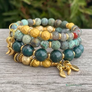 Puede incluir: Conjunto de pulseras de cuentas con cuentas de piedras preciosas verdes, doradas y grises. Se ven detalles y abalorios dorados. Las pulseras están apiladas sobre una superficie de madera. La palabra "BlooMoon" está en la parte inferior derecha.