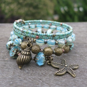 May include: A stack of beaded bracelets with turquoise, blue, and bronze accents. The bracelets feature glass beads, small seed beads, and bronze charms, including a seashell and starfish. The bracelets are on a wooden surface.