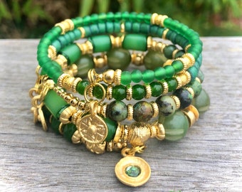 May include: A collection of green beaded bracelets with gold-coloured accents and charms. The bracelets feature various shades of green beads, including round, cylindrical, and textured styles. Gold-coloured metal spacers and charms add detail.