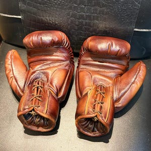 Puede incluir: Un par de guantes de boxeo de cuero marrón. Los guantes están atados por delante y tienen un aspecto vintage. El cuero tiene un color marrón rojizo intenso con sombras más oscuras.