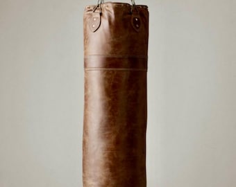 May include: A brown leather punching bag suspended from a chain. The cylindrical bag has a textured surface and a horizontal band. The top features a drawstring closure and is attached to a metal chain. It is designed for boxing and fitness training, a classic piece of equipment.