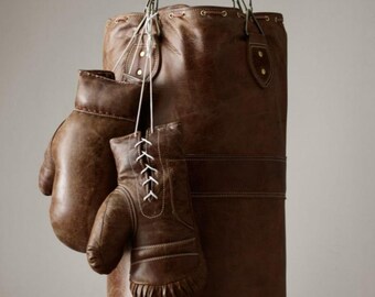 May include: A brown leather punching bag hangs from a chain, with a pair of matching boxing gloves suspended in front. The bag is cylindrical, with a drawstring closure at the top. The gloves are laced with white laces.