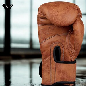 May include: A close-up of a brown leather boxing glove. The glove has a textured surface with visible stitching and small ventilation holes. The wrist strap and the area around the hand opening are black. The background is blurred.