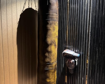 May include: A worn, brown and yellow leather punching bag suspended by a chain. Boxing gloves in black, brown, and tan are on the floor. A pair of boxing gloves are mounted on the wall. The scene is set in a gym.
