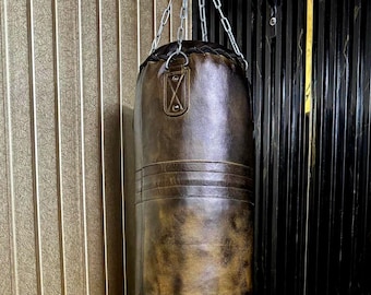 May include: A brown leather punching bag suspended from a silver chain. The bag has a distressed finish with horizontal stitched bands and a decorative emblem. The background features vertical wood paneling and a black wall.