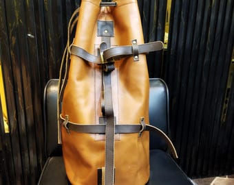Könnte beinhalten: Ein brauner Leder-Rucksack mit Kordelzug, dunklen braunen Riemen und Schnallen. Die Tasche ist zylindrisch und steht auf einem schwarzen Stuhl. Das Leder hat eine glatte, polierte Oberfläche, und die Riemen sind fest verschlossen.