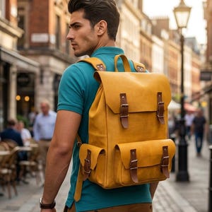 Può includere: Uno zaino in pelle color giallo senape dotato di un ampio scomparto principale con patta, due tasche frontali e cinturini in pelle marrone con fibbie in metallo. Lo zaino presenta spallacci regolabili e una maniglia superiore per il trasporto.
