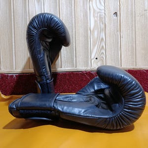 May include: A pair of black leather boxing gloves. The gloves are lying on a yellow surface.