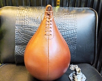 May include: A brown leather teardrop-shaped speed bag with a stitched seam down the center and a loop at the top. A silver metal swivel mount is next to the bag. The bag is designed for boxing training.