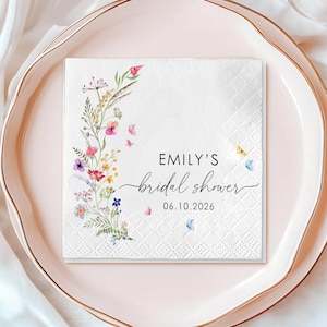 May include: A white square napkin with a floral design and the text "EMILY'S bridal shower 06.10.2026" is placed on a pink plate with a gold rim. The napkin features colorful flowers and butterflies.