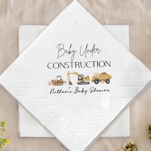 Op de afbeelding: Witte papieren servetten met een ruitpatroon. De bovenste servet heeft de tekst "Baby Under Construction" en "Nathan's Baby Shower" met illustraties van gele en bruine bouwmachines.