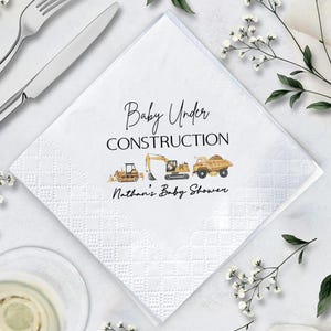 Op de afbeelding: Witte vierkante servet met een ruitpatroon en de tekst "Baby Under Construction" in zwart schrift. Daaronder illustraties van bouwmachines. De tekst "Nathan's Baby Shower" staat eronder.
