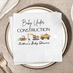 Op de afbeelding: Witte papieren servetten met een ruitpatroon, met de tekst "Baby Under Construction" en illustraties van bouwmachines. De servetten zijn voor Nathan's Baby Shower.