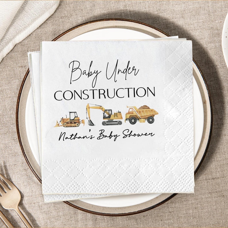 Op de afbeelding: Witte papieren servetten met een ruitpatroon, met de tekst "Baby Under Construction" en illustraties van bouwmachines. De servetten zijn voor Nathan's Baby Shower.