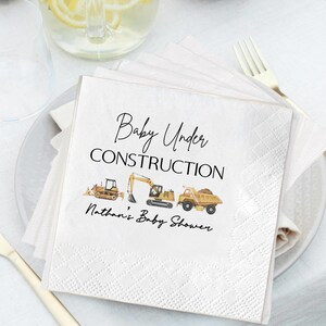 Op de afbeelding: Witte vierkante servetten met de tekst "Baby Under Construction" en "Nathan's Baby Shower" in zwart schrift. De servetten tonen illustraties van bouwmachines: een bulldozer, een graafmachine en een dumper. De servetten zijn op een wit bord gestapeld.