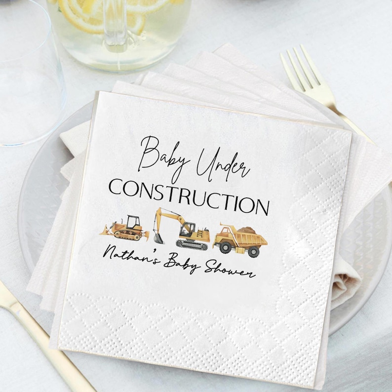 Op de afbeelding: Witte vierkante servetten met de tekst "Baby Under Construction" en "Nathan's Baby Shower" in zwart schrift. De servetten tonen illustraties van bouwmachines: een bulldozer, een graafmachine en een dumper. De servetten zijn op een wit bord gestapeld.